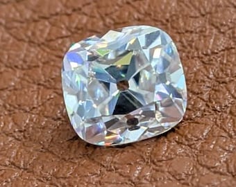 May include: A cushion-cut diamond with a brilliant, multifaceted surface. The gemstone is clear and reflects light, set against a brown leather background. The diamond's shape is square with rounded corners, showcasing its sparkle.