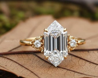 May include: A gold engagement ring with a large, emerald-cut diamond as the centerpiece. The ring features smaller diamonds on each side of the main stone and ornate detailing on the band. The ring is set on a brown leaf.