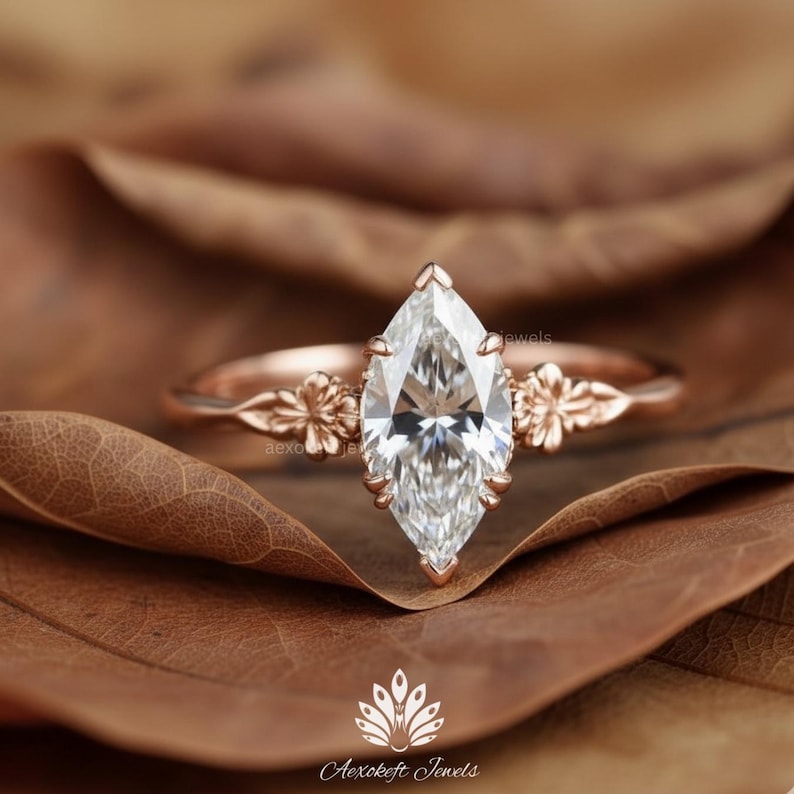 Puede incluir: Anillo de compromiso de oro rosa con un diamante talla marquesa. El anillo tiene detalles florales en la banda y alrededor del engaste del diamante. El anillo est&aacute; sobre una hoja marr&oacute;n.
