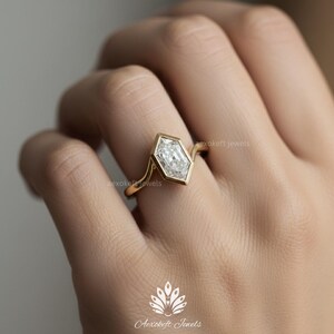May include: A gold ring with a unique, geometric-shaped diamond. The diamond is set in a bezel setting, and the band has a delicate, curved design. The ring is on a person's finger, showcasing the jewelry's elegant style.