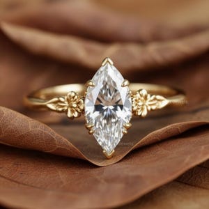 Puede incluir: Anillo de compromiso con diamante talla marquesa y banda dorada. El anillo presenta un diamante grande y transparente engastado en una montura de garras. La banda tiene detalles florales a cada lado del diamante. El anillo se muestra sobre una hoja marr&oacute;n.