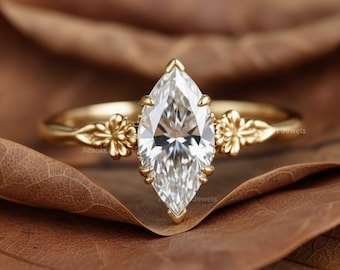 May include: A marquise-cut diamond engagement ring with a gold band. The ring features a large, clear diamond set in a prong setting. The band has floral accents on each side of the diamond. The ring is displayed on a brown leaf.