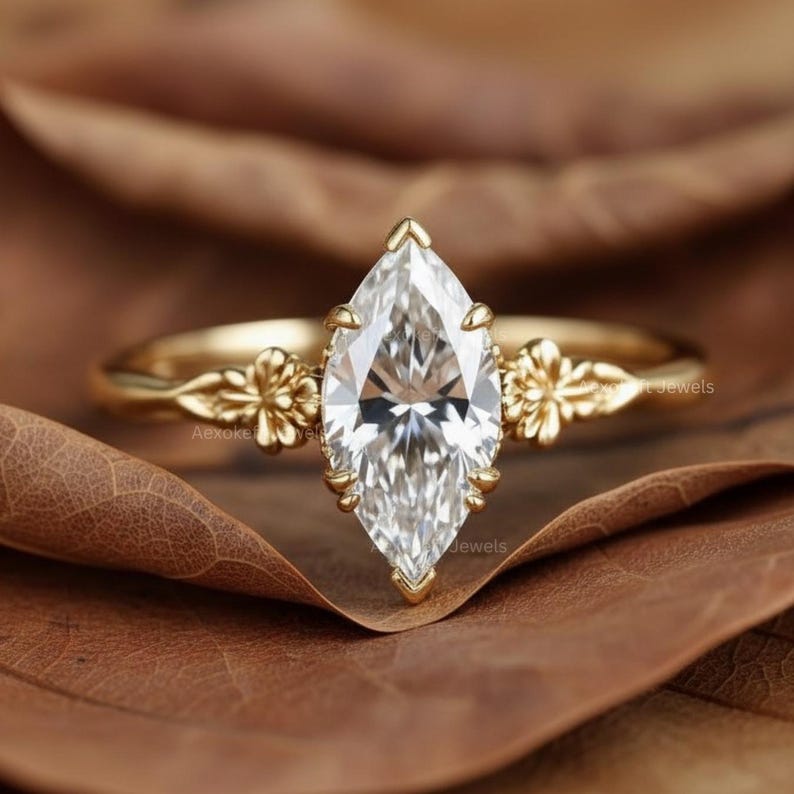 Puede incluir: Anillo de compromiso con diamante talla marquesa y banda dorada. El anillo presenta un diamante grande y transparente engastado en una montura de garras. La banda tiene detalles florales a cada lado del diamante. El anillo se muestra sobre una hoja marr&oacute;n.