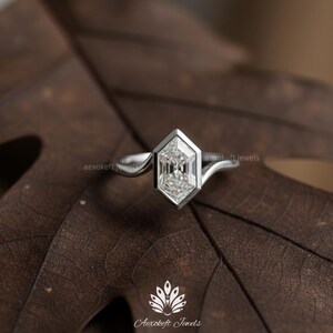 May include: A silver ring with a geometric, hexagonal-shaped diamond. The diamond is set in a bezel setting. The ring is on a brown leaf. The brand name "Aexokeft Jewels" is visible.