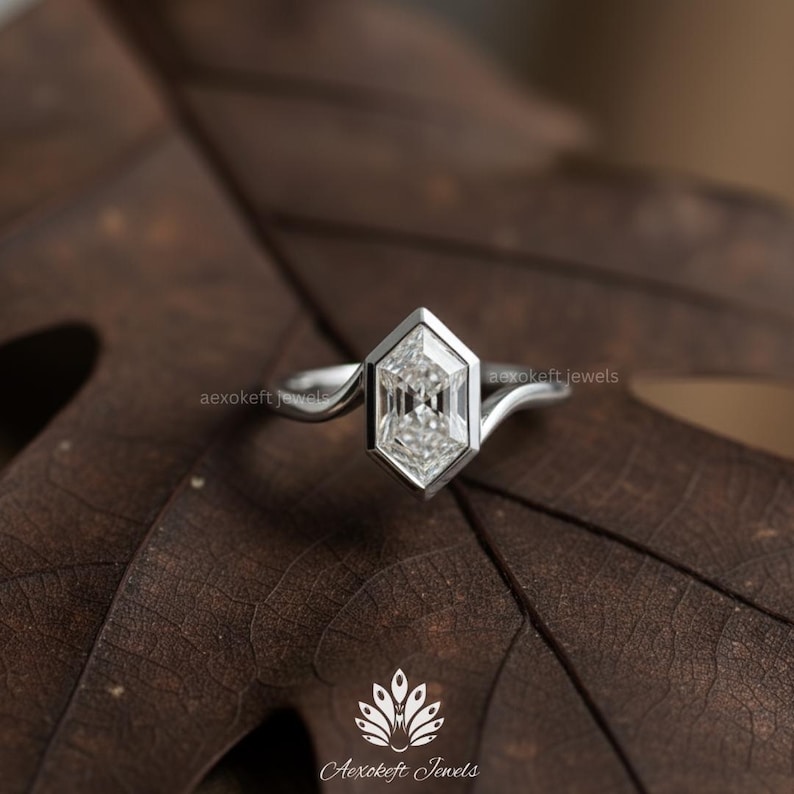 May include: A silver ring with a geometric, hexagonal-shaped diamond. The diamond is set in a bezel setting. The ring is on a brown leaf. The brand name "Aexokeft Jewels" is visible.