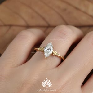 Puede incluir: Un anillo de oro con un diamante talla marquesa, flanqueado por peque&ntilde;os detalles florales. El anillo tiene una banda delicada. El anillo es una joya. El fondo es neutro.