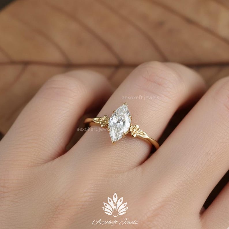 Puede incluir: Un anillo de oro con un diamante talla marquesa, flanqueado por peque&ntilde;os detalles florales. El anillo tiene una banda delicada. El anillo es una joya. El fondo es neutro.