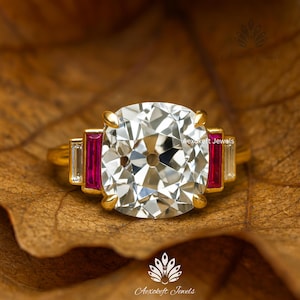 Puede incluir: Un anillo de oro con un gran diamante transparente de talla cojín como pieza central. El anillo está flanqueado por piedras rectangulares rojas y blancas. El anillo está sobre una hoja marrón.
