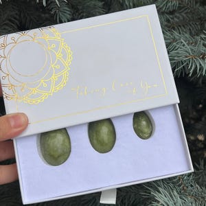 May include: A white box with gold accents and the text "Taking Care of You" contains three green, oval-shaped stones. The box has a gold mandala design on the top left corner. The stones are nestled in a white interior.
