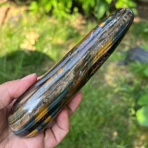 Puede incluir: Una piedra pulida y alargada con una mezcla de tonos azules, dorados y marrones. La piedra tiene una superficie lisa y brillante y una forma redondeada. Los colores están arremolinados y en capas, creando un patrón natural y orgánico. La piedra se sostiene en una mano sobre un fondo verde borroso.