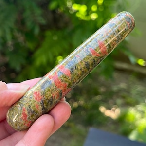 Puede incluir: Una varita de cristal de unakita pulida. La varita es cilíndrica con extremos redondeados. Presenta un patrón moteado de verde, rosa y negro. El cristal se sostiene en una mano sobre un fondo verde borroso.