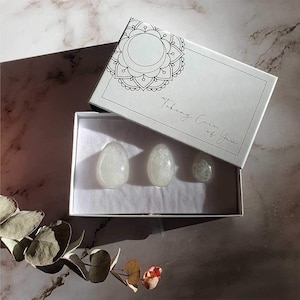 May include: A white gift box with the text "Taking Care of You" contains three smooth, oval-shaped stones. The box lid features a decorative circular design. The stones are arranged on a white surface, with a marble background.