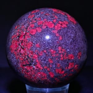 Può includere: Una sfera di pietra preziosa lucidata, con un colore di base viola scuro, punteggiata da vivaci macchie rosse. La superficie lucida riflette la luce, creando un effetto luminoso. La sfera è esposta su un supporto trasparente.