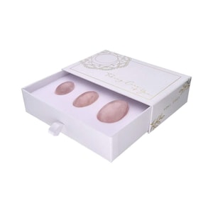 May include: A white box with a drawer containing three pink, oval-shaped stones. The box has gold floral accents and text that reads "Taking Care of You" and "Yoni Eggs". The drawer is open, revealing the stones nestled in a white interior.