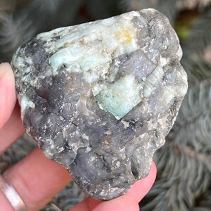 Peut inclure: Une émeraude brute, mélangeant des teintes grises, vertes et blanches. La pierre naturelle présente une surface texturée avec des structures cristallines visibles. L'émeraude est tenue dans une main, révélant sa forme brute et non polie.