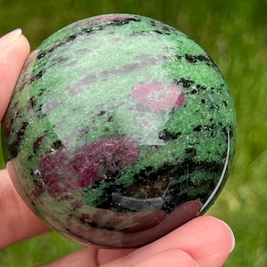 May include: A green, pink, and black gemstone sphere. The sphere is polished and has a smooth surface.