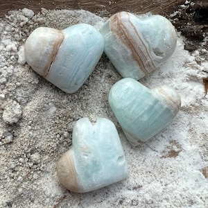 May include: Four heart-shaped blue and white calcite crystal stones. The crystals have a natural, rough texture and are arranged on a white surface.