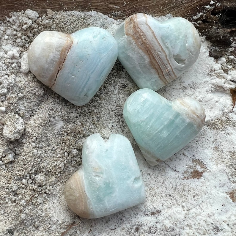 May include: Four heart-shaped blue and white calcite crystal stones. The crystals have a natural, rough texture and are arranged on a white surface.