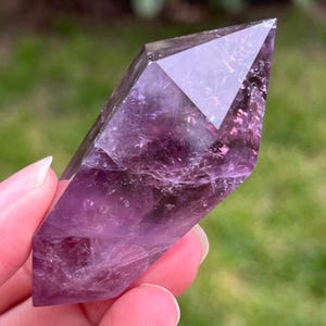 May include: A faceted amethyst crystal point, showcasing a gradient of deep purple hues. The crystal's polished facets reflect light, creating a shimmering effect. The crystal is held against a blurred green background.