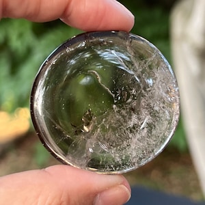 Può includere: Una sfera di cristallo di quarzo trasparente con una superficie naturale e ruvida e un piccolo pezzo di pirite all'interno. La sfera ha un diametro di circa 5 cm.