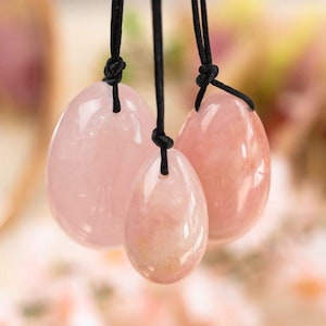 May include: Three rose quartz eggs of varying sizes, each suspended by a black cord. The eggs have a smooth, polished surface and a soft pink hue. The background is blurred, with hints of pink and green.