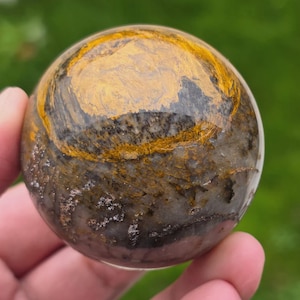 Può includere: Una gemma sferica lucidata con un motivo a spirale di tonalità oro, grigio e marrone. L'orbita è tenuta in una mano, mostrando la sua superficie liscia e riflettente. Le variazioni di colore della pietra creano un display accattivante.