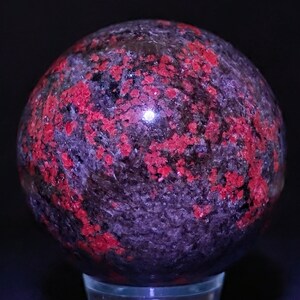 May include: A polished sphere with a dark purple base color, speckled with vibrant red patches. The sphere is smooth and reflective, with a glossy finish. The overall effect is a striking contrast of colors and textures.