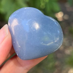 Könnte beinhalten: Ein herzförmiger Stein in einem sanften, gedämpften Blau. Die polierte Oberfläche reflektiert das Licht und hebt subtile Farbvariationen hervor. Der Stein wird in einer Hand gehalten, mit einem verschwommenen grünen Hintergrund.