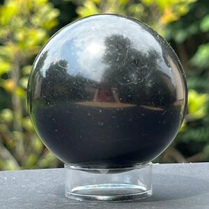 Könnte beinhalten: Eine schwarze Obsidiankugel auf einem klaren Acrylsockel. Die Kugel ist poliert und reflektiert Licht.