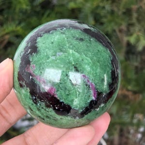 Op de afbeelding: Een gepolijste bol van groene zoisiet met zwarte en robijn insluitsels. De edelsteen wordt in een hand gehouden, waarbij het gladde, reflecterende oppervlak wordt getoond. De kleuren en patronen van de bol creëren een opvallend visueel effect.