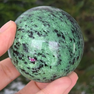 Op de afbeelding: Een gepolijste bol van groene zoisiet met zwarte en roze insluitsels. De edelsteen wordt in een hand gehouden, wat het gladde, reflecterende oppervlak laat zien. De bol heeft een levendige groene kleur met donkere vlekken en een klein roze gebied.