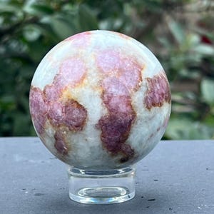 May include: A white, pink, and brown crystal sphere on a clear acrylic stand. The sphere has a swirling pattern of pink and brown.