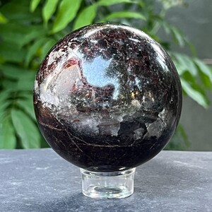 Peut inclure: Une sphère polie, rouge foncé, avec des surfaces réfléchissantes, posée sur un socle en verre transparent. La sphère présente un aspect marbré avec des inclusions cristallines plus claires. Fond vert flou.