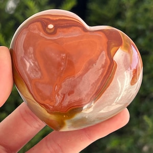 May include: A heart-shaped polished stone in shades of orange, brown, and cream. The stone has a smooth, glossy finish and is held in a hand. The pattern on the stone features swirling bands of colour.