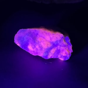 May include: A fluorescent rock, lit by ultraviolet light, displaying a mix of purple, pink, and orange colours. The rock has an irregular shape and a rough texture, set against a dark background.