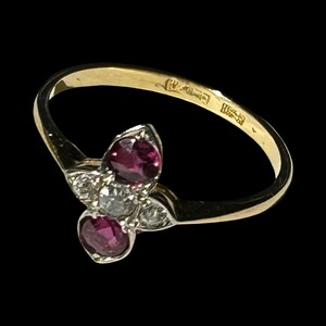 May include: A gold ring with a cluster of gemstones. The ring features two oval-cut, deep red rubies and three small, clear diamonds. The band is a simple, polished gold. The ring is set against a black background.