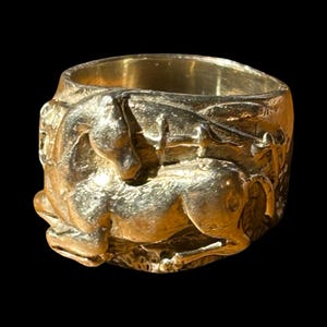Puede incluir: Un anillo dorado con un relieve detallado de un caballo. El anillo tiene una banda ancha y una superficie texturizada, con el caballo representado en posición reclinada. El fondo es negro.