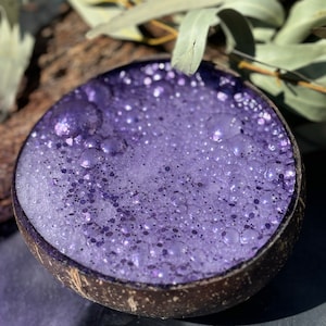 May include: A purple, foamy drink in a coconut shell. The drink is topped with a layer of bubbles.
