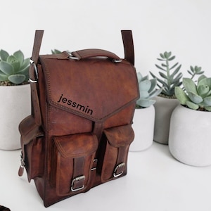 May include: A brown leather backpack featuring a top handle, two vertical front pockets with buckle closures, and side pockets. The word jessmin is printed in black on the front flap. The bag has adjustable shoulder straps and metal hardware.
