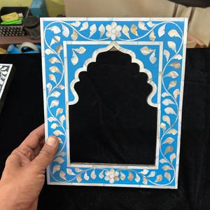 May include: A square, light blue picture frame with a central arch design. The frame is decorated with white floral and leaf patterns, and mother-of-pearl accents. The frame is held by a hand.