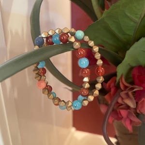 May include: Two beaded bracelets with gold, turquoise, coral, and red beads. The bracelets are stacked on top of each other.