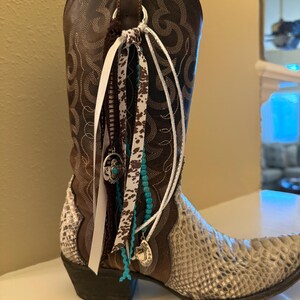 Puede incluir: Una bota vaquera de cuero marrón con pie de textura de piel de serpiente, costuras intrincadas en la caña y un accesorio decorativo unido a la trabilla, compuesto por cintas blancas, marrones y turquesas, cuentas y colgantes de metal.