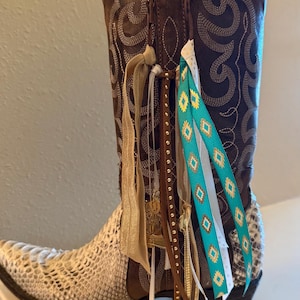 Puede incluir: Una bota de vaquero de cuero marrón con pie de piel de serpiente y costuras decorativas. La bota está adornada con cintas colgantes en turquesa, blanco y dorado, junto con abalorios y un tirador de cremallera dorado. Un accesorio elegante de temática western.