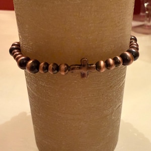 Op de afbeelding: Een koperkleurige kralenarmband met een kruisvormige sluiting. De armband is om een beige kaars gewikkeld. De kralen zijn donkerbruin en koperkleurig. De kruissluiting heeft een bijpassende koperkleur. De armband is een religieus accessoire.