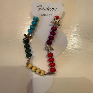 Op de afbeelding: Enkelbandje met kleurrijke kralen en gouden zeesterbedels. Het enkelbandje heeft een kleurengradatie, waaronder turquoise, groen, geel, rood en bordeaux. Het label leest "Fashion ANKLET".