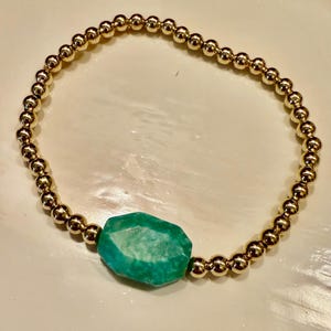 May include: A gold beaded bracelet with a single, large, green gemstone charm.