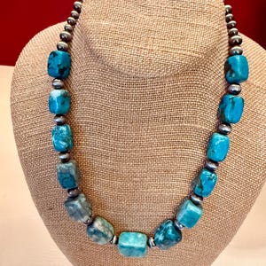 May include: A necklace featuring alternating polished turquoise stones and small metallic beads. The turquoise stones are rectangular with rounded edges, displaying shades of blue and green with dark veining. The metallic beads are round and silver-toned.