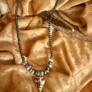 May include: A necklace with a gold arrowhead pendant and brown and blue beads. The necklace is on a brown velvet background.
