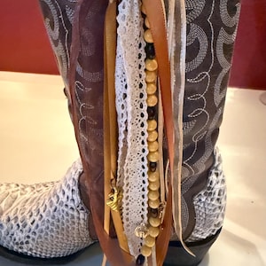 May include: A dark brown leather cowboy boot with white stitching and a textured white snakeskin-patterned foot. A decorative tassel hangs from the side, featuring cream lace, brown and tan ribbons, and a strand of round wooden beads with charms.