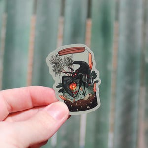 Peut inclure: Un autocollant représentant une créature noire aux yeux orange vif et au cœur flamboyant, enfermée dans un bocal en verre. L'illustration comprend des plantes et une base marron, avec des accents orange et verts. L'autocollant est tenu par une main.